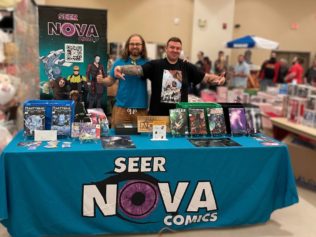 SeerNova Comics tabling at Saratoga Comic Con in 2024 with their comics displayed on a table wrapped in the SeerNova Comics Logo and a banner behind them with the logo and characters as well as a QR Code.